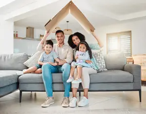 A family of four, two adults and two children, are sitting on a couch in a living room and posing for a photo with a cardboard house above their heads.