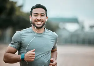 A man wearing earphones and a gray t-shirt is smiling and running on a road during the day