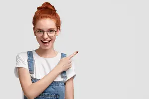 red-haired woman wearing glasses and denim overalls pointing finger at something