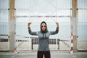 A woman in a gray hoodie and sunglasses poses in front of a metal gate near a beach.