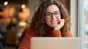 A smiling woman with curly hair and glasses, sitting in front of a laptop in a coffee shop.