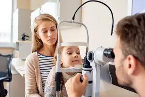 A young girl is having her eyes checked by a doctor with a woman watching