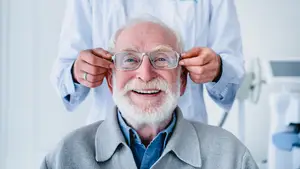 An elderly man with a white beard and mustache wearing glasses is being examined by a doctor in a white coat, holding his glasses and smiling with his eyes closed in a white room.
