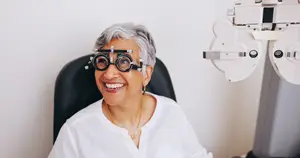 A woman with gray hair is smiling and wearing a white shirt and glasses, she is sitting in a chair in front of an eye examination machine, which is on the right side, and she is wearing a necklace.