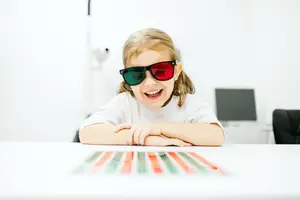 A girl with sunglasses is smiling and leaning on a table with colored pencils on it.