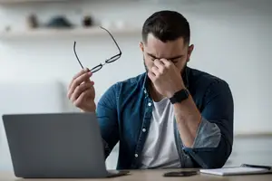 Businessman holding glasses with tired eyes looking at a laptop