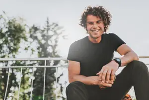 A man wearing a black t-shirt and black pants sitting on a metal fence and smiling with curly hair and wearing a watch.