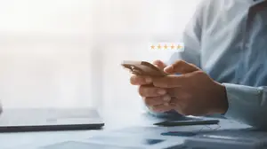 A man is using a smartphone with a five-star rating icon on the screen while sitting at a desk with a laptop and a pen.