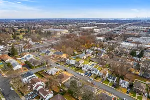 An aerial view of a suburban neighborhood with houses, a road, and a highway in the distance under a blue sky with clouds