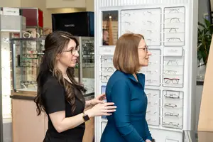 Two women wearing glasses standing in front of an optical shop displaying various frames.