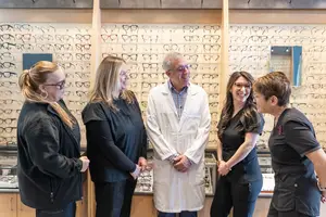 Five women and a man standing in front of a display of glasses in an optical shop, all smiling and looking at the camera
