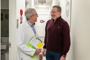 A smiling male doctor wearing a white coat and a male patient wearing a maroon sweater and a watch stand in a hallway and talk to each other