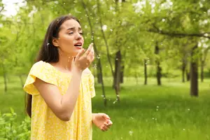 A woman standing in a grassy field with trees behind her, she is blowing bubbles with her mouth and her eyes are closed. She is wearing a yellow dress with a white pattern.
