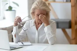 Elderly woman sitting at a desk, covering her eyes with her hand, perhaps feeling stressed or overwhelmed