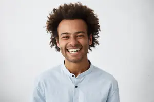 A young man with a wide smile on his face is standing in front of a white wall, wearing a blue shirt and black buttons.