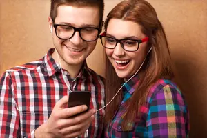 A young man and woman with headphones on and smiling, listening to music on their phone.