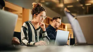 A woman wearing glasses and a striped shirt is sitting at a table with a laptop in front of her, and a man is sitting behind her with a laptop in front of him. They are sitting in a room with a wooden cabinet behind them.