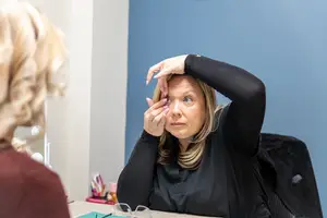 A woman wearing a black shirt is sitting in front of a mirror and appears to be putting in a contact lens