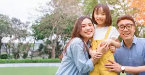 A family of three smiling and posing for a photo in a park with trees and a building in the background