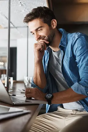 A man sitting at a desk with a laptop, smiling while wearing a denim shirt and a wristwatch.