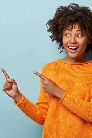 A young woman with curly hair, wearing an orange sweater, is smiling and pointing her index fingers at an unseen object in front of her.