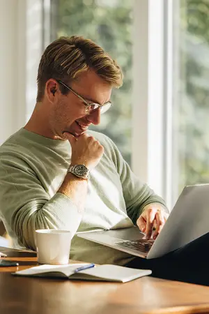 A man is sitting on a chair while smiling and typing on a laptop on a table
