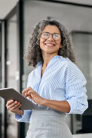 A smiling woman wearing glasses and a blue and white striped shirt, holding a tablet and standing in an office with glass walls.