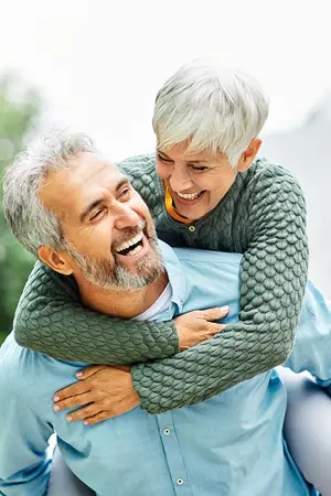 An elderly man with white hair and beard is hugging an elderly woman with white hair and a green sweater. The elderly woman is smiling and hugging the elderly man. They are both looking at each other. Behind them is a blurry background of trees and a white sky.