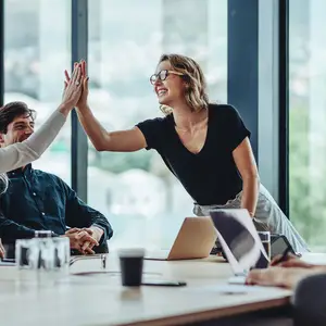 A woman wearing glasses is high-fiving a man sitting at a table with laptops and cups