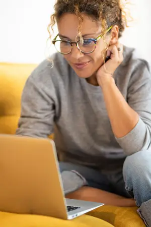 A woman with glasses is sitting on a couch and seems to be talking on a cellphone while looking at her laptop.