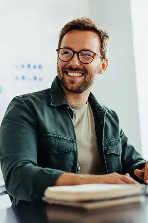 A smiling man with glasses sitting at a desk and looking at a book