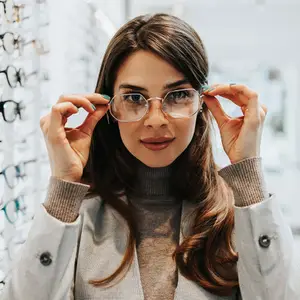 A woman wearing eyeglasses is standing in front of a rack of glasses and is holding the frames in her hands, possibly posing for a photo.