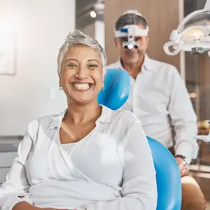 Woman sitting in a dental chair while a male dentist is examining her teeth with dental tools and a light.