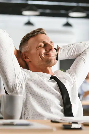 businessman sitting at desk in office with a cup and papers smiling with his arms behind his head