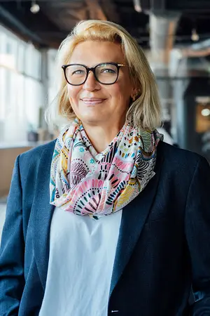 A smiling woman with blonde hair and glasses standing in a modern office with white walls and glass windows. She is wearing a blue blazer, a white shirt, and a colorful scarf around her neck. Behind her, there is a white shelf with some items on it.