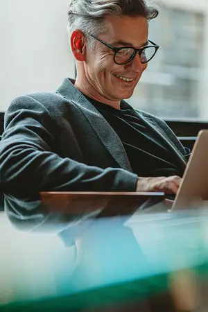 A man wearing glasses is smiling while working on a laptop on a glass table in an office.