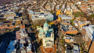 An aerial view of a city with numerous buildings, including a large cathedral-like structure with a dome and spire, surrounded by other buildings of various sizes and styles, all under a clear sky.