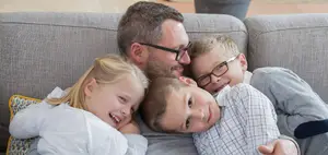A man and his three children laying on a couch together smiling at the camera.