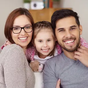 A family of three, including a woman, a man, and a girl, is posing for a photo with smiles on their faces, possibly inside a house.