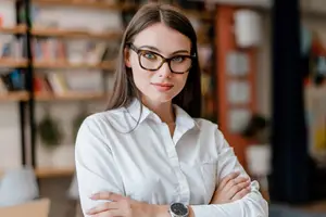 A woman wearing glasses and a white shirt stands in an office with her arms crossed