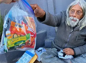 An elderly man with white hair and beard is sitting on the ground holding a bag of food with a label that reads Hot Hands and Toe Warmers.