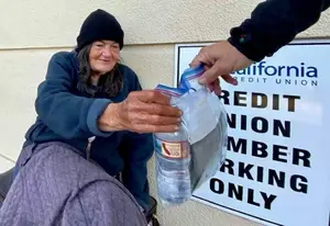 A woman in a blue jacket and black beanie sits next to a sign for California Credit Union, holding a plastic bag with a bottle inside, as a person hands her a bag with a zipper.