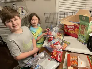 Two children are standing next to a table with a variety of food items in plastic bags, boxes, and containers, one of them smiling, while the other is looking at the camera.