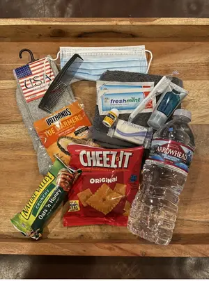 A collection of items on a wooden surface including a bottle of Arrowhead water, a pack of Cheez-It crackers, a bag of HotHands warmers, a pair of socks, a bottle of toothpaste, a comb, a face mask, and an American flag pin.