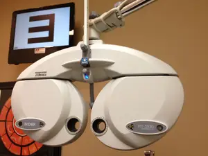 An eye testing machine with a screen behind it, displaying a letter E, is mounted on a wall. It is a NIDEK RT-5100 refractor.