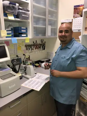A man in a blue shirt is standing behind a desk in a lab with various equipment and papers on the desk.