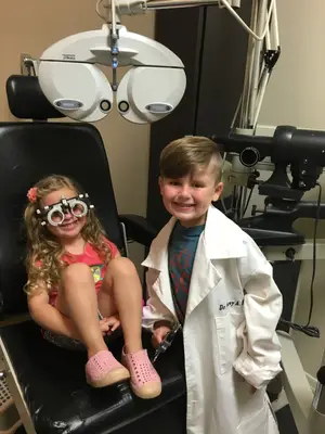 Two children smiling with one sitting in an optometrist chair and the other wearing a white lab coat