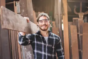 A man wearing glasses and a plaid shirt stands in a workshop holding a large wooden plank