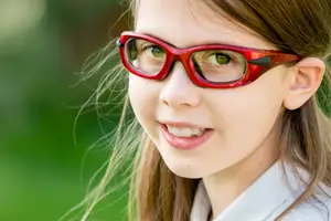 A girl with long brown hair and red glasses smiling for a picture in a park