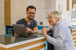 A man and an elderly woman are smiling and looking at a laptop at a reception desk